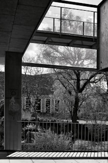 A black and white photograph featuring an urban outdoor scene with a modern architectural structure in the foreground. A tree with bare branches is visible in the background, casting intricate shadows on the ground. A railing with vertical bars is prominently positioned, with sunlight casting linear patterns. Graffiti, including a mushroom and the words 'Happy Life,' is painted on a pillar.
