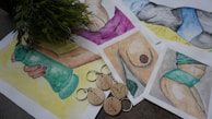 The image features several watercolor paintings depicting various body parts and fitness themes, including a hand holding a green dumbbell, a torso, and a leg with a tattoo. Some artworks involve abstract representations, and all are arranged on a surface. A bunch of green leaves partially covers part of the artworks. Wooden keychains with minimalist line art designs are placed over the paintings.
