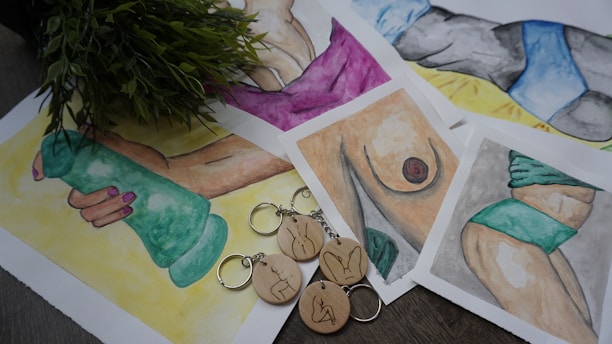 The image features several watercolor paintings depicting various body parts and fitness themes, including a hand holding a green dumbbell, a torso, and a leg with a tattoo. Some artworks involve abstract representations, and all are arranged on a surface. A bunch of green leaves partially covers part of the artworks. Wooden keychains with minimalist line art designs are placed over the paintings.