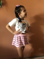 A young girl with a cheerful expression, wearing a white t-shirt with zebra patterns and a pink skirt with black stripe designs. Her hair is tied back with a colorful hair tie, and she stands against a plain orange wall, laughing.