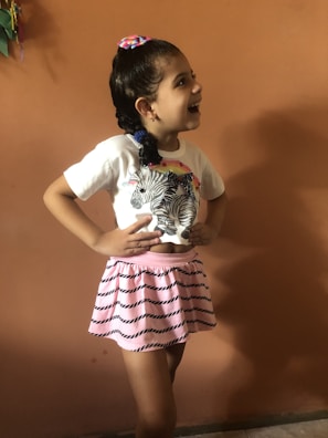 A young girl with a cheerful expression, wearing a white t-shirt with zebra patterns and a pink skirt with black stripe designs. Her hair is tied back with a colorful hair tie, and she stands against a plain orange wall, laughing.
