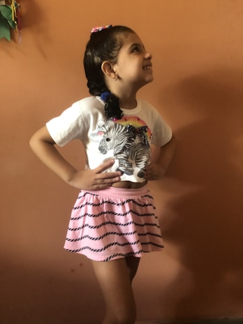 A young girl is smiling and looking upwards with her hands on her hips. She is wearing a white t-shirt with a zebra print design and a pink skirt with black stripes. Her hair is styled in braids with colorful hair ties. The background is a plain orange wall.