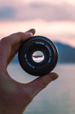 A hand holds a Fujinon aspherical lens with a scenic landscape visible through the lens opening. The background is softly blurred, featuring subtle colors that suggest a sunset over water or mountains.