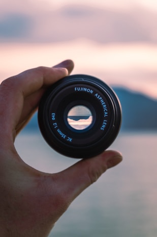 A hand holds a Fujinon aspherical lens with a scenic landscape visible through the lens opening. The background is softly blurred, featuring subtle colors that suggest a sunset over water or mountains.