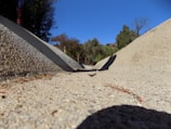 A dry concrete drainage ditch slopes downward, bordered by leafy trees under a clear blue sky. The foreground shows the texture and details of the concrete surface.