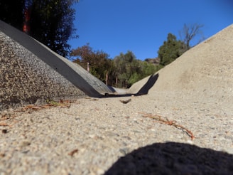 A dry concrete drainage ditch slopes downward, bordered by leafy trees under a clear blue sky. The foreground shows the texture and details of the concrete surface.