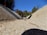 A dry concrete drainage ditch slopes downward, bordered by leafy trees under a clear blue sky. The foreground shows the texture and details of the concrete surface.