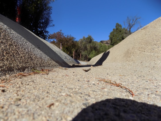 A dry concrete drainage ditch slopes downward, bordered by leafy trees under a clear blue sky. The foreground shows the texture and details of the concrete surface.