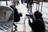 A person in dark clothing is sitting on a high chair, holding a large camera, and appears to be filming or photographing another individual who is seated at a table with a mosaic design. Another person is walking in the background on a paved outdoor area with scattered tables and chairs.