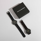 A stylish watch box labeled 'Chikiboom' is positioned next to a pair of minimalist wristwatches. Both watches have dark bands and simple, elegant clock faces with thin hands and markers.