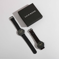 A stylish watch box labeled 'Chikiboom' is positioned next to a pair of minimalist wristwatches. Both watches have dark bands and simple, elegant clock faces with thin hands and markers.