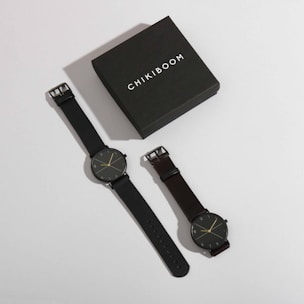 A stylish watch box labeled 'Chikiboom' is positioned next to a pair of minimalist wristwatches. Both watches have dark bands and simple, elegant clock faces with thin hands and markers.