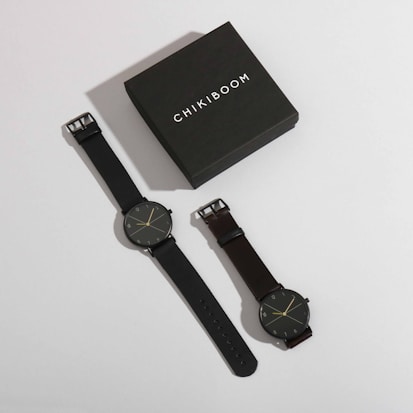 A stylish watch box labeled 'Chikiboom' is positioned next to a pair of minimalist wristwatches. Both watches have dark bands and simple, elegant clock faces with thin hands and markers.