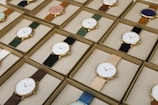 A collection of boxed wristwatches neatly arranged in rows, each featuring a minimalist design with a variety of strap colors. The watches have round faces with simple markers and are encased in what appears to be gold or golden metal, adding a touch of elegance.