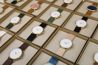 A collection of boxed wristwatches neatly arranged in rows, each featuring a minimalist design with a variety of strap colors. The watches have round faces with simple markers and are encased in what appears to be gold or golden metal, adding a touch of elegance.