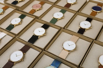 A collection of boxed wristwatches neatly arranged in rows, each featuring a minimalist design with a variety of strap colors. The watches have round faces with simple markers and are encased in what appears to be gold or golden metal, adding a touch of elegance.
