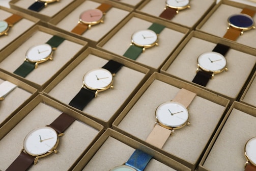 A collection of boxed wristwatches neatly arranged in rows, each featuring a minimalist design with a variety of strap colors. The watches have round faces with simple markers and are encased in what appears to be gold or golden metal, adding a touch of elegance.