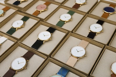 A collection of boxed wristwatches neatly arranged in rows, each featuring a minimalist design with a variety of strap colors. The watches have round faces with simple markers and are encased in what appears to be gold or golden metal, adding a touch of elegance.