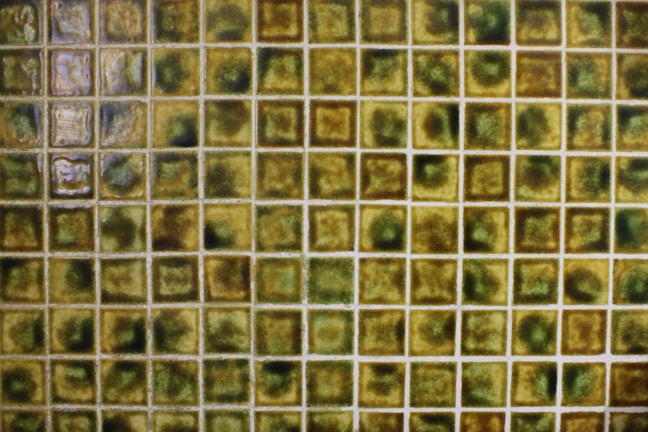 A grid of small, square, ceramic tiles featuring a textured surface. The tiles display a variety of earthy tones, including shades of green, brown, and yellow, with occasional glossy highlights from reflections.