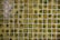 A grid of small, square, ceramic tiles featuring a textured surface. The tiles display a variety of earthy tones, including shades of green, brown, and yellow, with occasional glossy highlights from reflections.