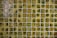 A grid of small, square, ceramic tiles featuring a textured surface. The tiles display a variety of earthy tones, including shades of green, brown, and yellow, with occasional glossy highlights from reflections.