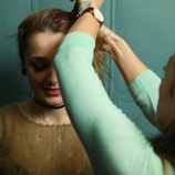A woman is having her hair styled by another person whose arms are visible. The background is a teal-colored wall, and the woman has light brown hair, red lipstick, and a choker necklace. She is wearing a brown, patterned top. The stylist is wearing a watch and has a light green sleeve.
