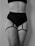 A black and white image featuring a person wearing a black high-waisted undergarment with adjustable straps attached to garters on the thighs. The focus is on the midsection and thighs, with no visible facial features.