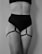 A black and white image featuring a person wearing a black high-waisted undergarment with adjustable straps attached to garters on the thighs. The focus is on the midsection and thighs, with no visible facial features.
