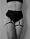 A black and white image featuring a person wearing a black high-waisted undergarment with adjustable straps attached to garters on the thighs. The focus is on the midsection and thighs, with no visible facial features.