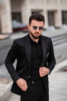 A man is wearing a tailored black suit with a matching black shirt and stylish sunglasses. He has a well-groomed beard and is standing in an urban setting. The background is slightly blurred, suggesting a street or modern building facade.