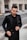 A man is wearing a tailored black suit with a matching black shirt and stylish sunglasses. He has a well-groomed beard and is standing in an urban setting. The background is slightly blurred, suggesting a street or modern building facade.