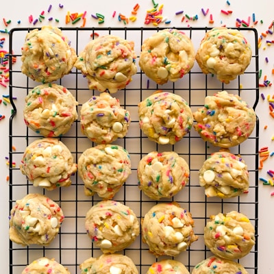 A batch of colorful cookies with white chocolate chips and rainbow sprinkles rests on a black cooling rack. The cookies are arranged neatly in rows, with loose sprinkles scattered around.
