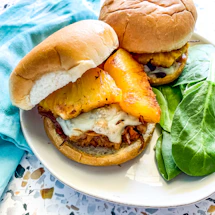Two sandwiches are served on a white plate. Each sandwich is in a soft bun with a slice of grilled pineapple, melted cheese, and what appears to be pulled pork. Fresh spinach leaves are placed alongside as garnish. A turquoise cloth lies next to the plate on a terrazzo countertop.