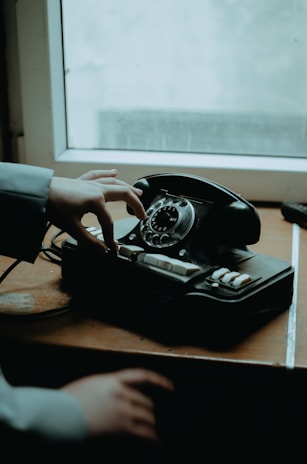 A rotary dial telephone is placed on a wooden surface near a window. A person's hand is reaching towards the dial of the vintage phone. The scene is dimly lit, suggesting an old-fashioned or nostalgic setting.