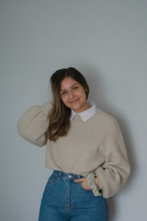 A woman stands against a plain background with a slight smile on her face. She is wearing a light beige sweater over a white collared shirt and high-waisted blue jeans. Her hair is styled to one side, and she holds a scrunchie in her left hand.