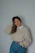 A smiling woman wearing a cozy beige sweater from Seeva Wear, standing against a soft chocolate brown background.