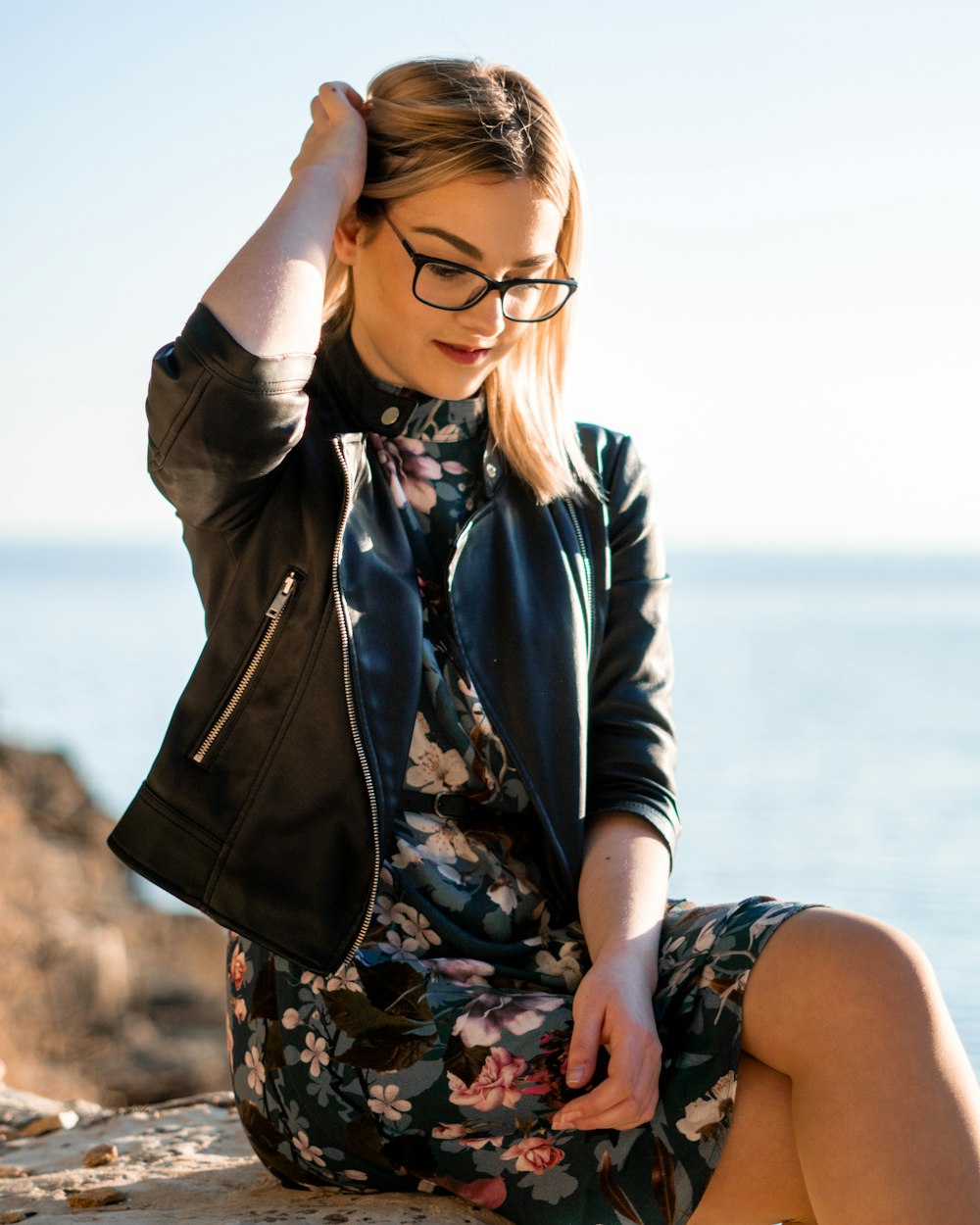 woman in black framed eyeglasses and brown jacket sitting on rock during daytime
