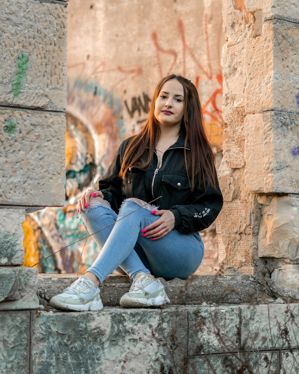 Frau in schwarzer Jacke und blauer Jeans sitzt auf Betonbank