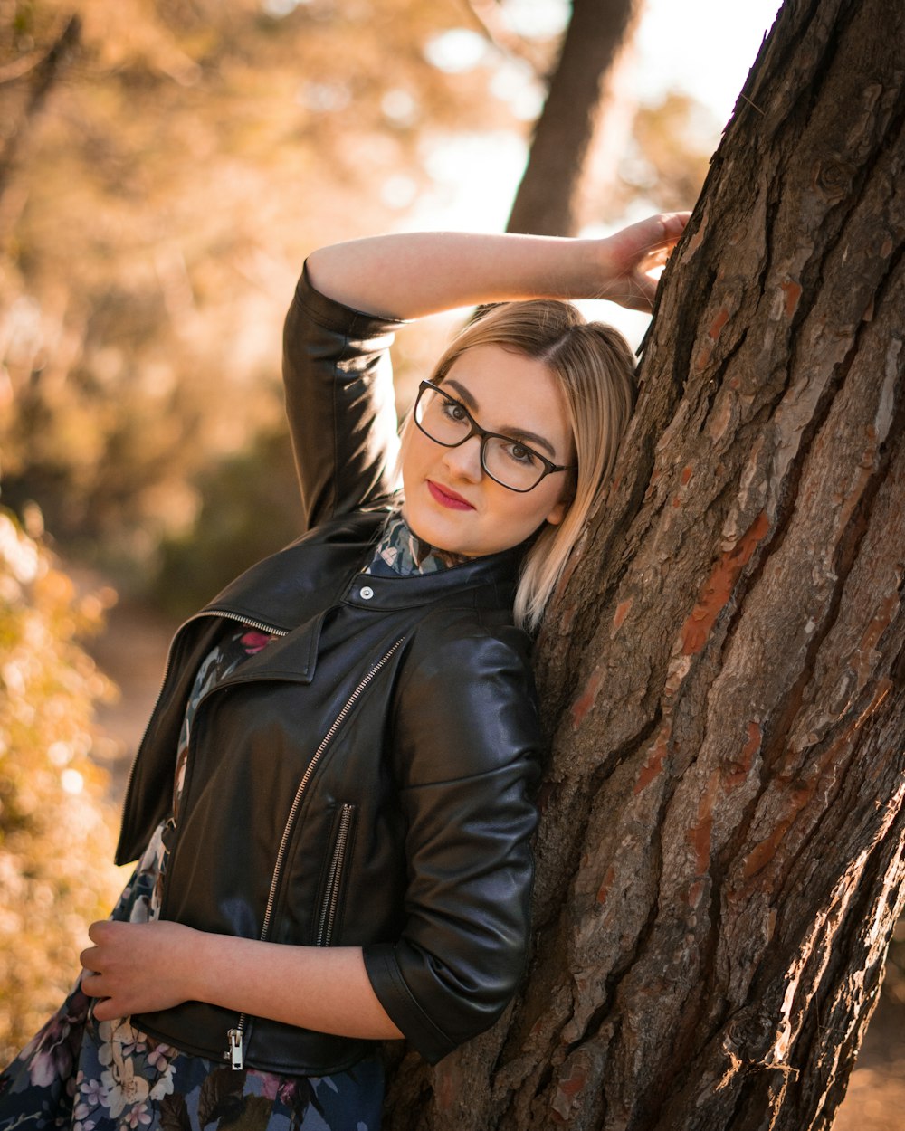woman in black leather jacket leaning on brown tree during daytime