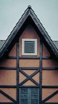 A traditional half-timbered house facade with a steeply pitched roof. Dark wooden beams form a crosshatch pattern against light brown walls. Two sets of shutters are visible, one on a small attic window and another larger one below.