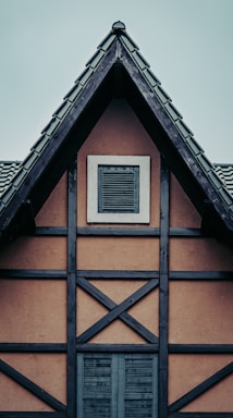 A traditional half-timbered house facade with a steeply pitched roof. Dark wooden beams form a crosshatch pattern against light brown walls. Two sets of shutters are visible, one on a small attic window and another larger one below.
