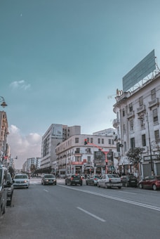 A city street lined with a variety of mid-rise buildings featuring shops and businesses. Several cars are stopped at a traffic light under a clear blue sky. The street is busy and well-maintained, with a few pedestrians visible. The architecture combines traditional and modern elements, and a large empty billboard is mounted on one of the buildings.