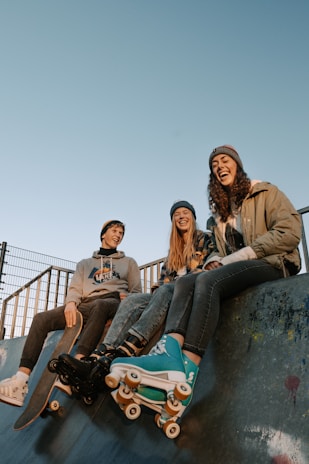 Three young people are sitting on the edge of a ramp at a skatepark. They are casually dressed, wearing beanies and jackets. One of them is holding a skateboard, while another is wearing rollerskates. They appear to be enjoying each other's company.