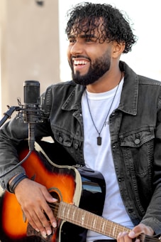 A man with curly hair and a beard is playing an acoustic guitar. He is wearing a denim jacket over a white T-shirt and a necklace with a small pendant. A microphone is positioned in front of him, suggesting he is performing or recording music. The background is out of focus, emphasizing the subject.