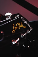 A close-up view of a guitar headstock featuring tuning pegs and the text 'Les Paul Model' inscribed in a stylish script. The lighting is subdued, with reflections highlighting the metallic parts and a warm glow on the text.