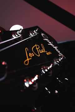 A close-up view of a guitar headstock featuring tuning pegs and the text 'Les Paul Model' inscribed in a stylish script. The lighting is subdued, with reflections highlighting the metallic parts and a warm glow on the text.