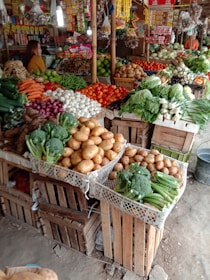 A market stall filled with a wide variety of fresh vegetables and produce. Potatoes, tomatoes, carrots, broccoli, onions, and garlic are neatly arranged in wooden crates. A person is standing behind the stall, organizing packaged goods hanging above, such as seasoning packets and dried products. The produce is displayed in a vibrant, colorful, and organized manner.