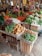 A market stall filled with a wide variety of fresh vegetables and produce. Potatoes, tomatoes, carrots, broccoli, onions, and garlic are neatly arranged in wooden crates. A person is standing behind the stall, organizing packaged goods hanging above, such as seasoning packets and dried products. The produce is displayed in a vibrant, colorful, and organized manner.