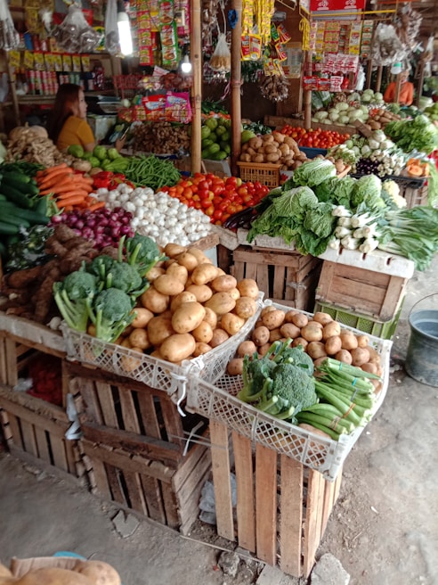A market stall filled with a wide variety of fresh vegetables and produce. Potatoes, tomatoes, carrots, broccoli, onions, and garlic are neatly arranged in wooden crates. A person is standing behind the stall, organizing packaged goods hanging above, such as seasoning packets and dried products. The produce is displayed in a vibrant, colorful, and organized manner.