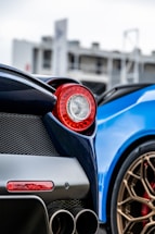 A close-up of a luxury sports car featuring a red tail light, dual exhaust pipes, and a portion of another car with a bright blue exterior and bronze wheels in the background. The focus is on the design elements and color contrast between the dark and vibrant hues.